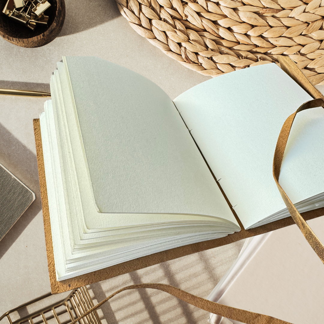 right photo of an open personalised leather journal placed next to a wicker coaster and binder clips, bathed in warm natural sunlight for a cozy creative workspace feel