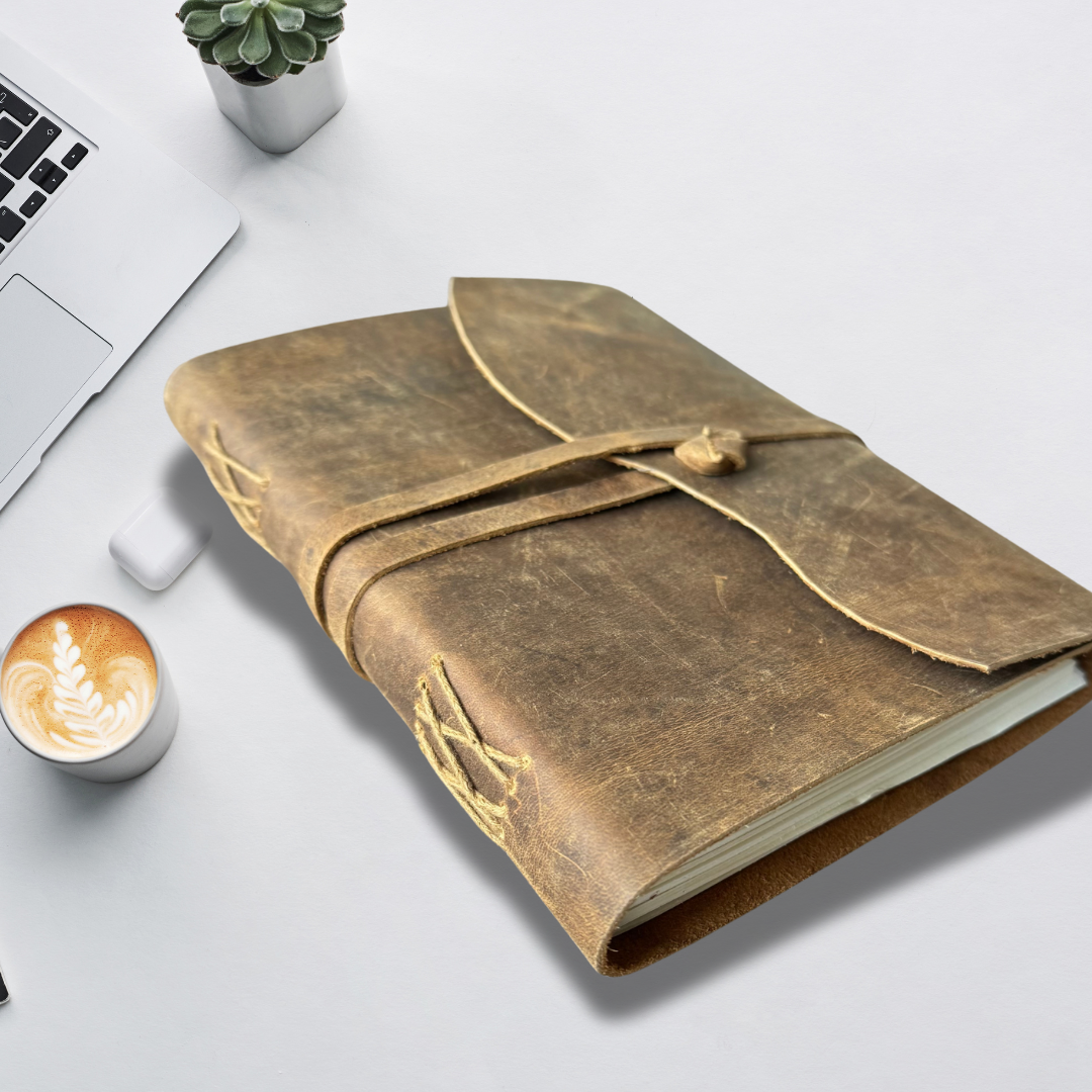 Photo of a personalised leather journal placed on a wooden desk alongside a laptop, steaming coffee cup, and a small green plant, creating a cozy and productive workspace vibe