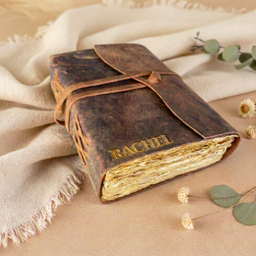 Handmade leather bound journal with gold embossed name RACHEL, styled on cream linen wrap with eucalyptus and dried florals — personalised leather journal gift Australia